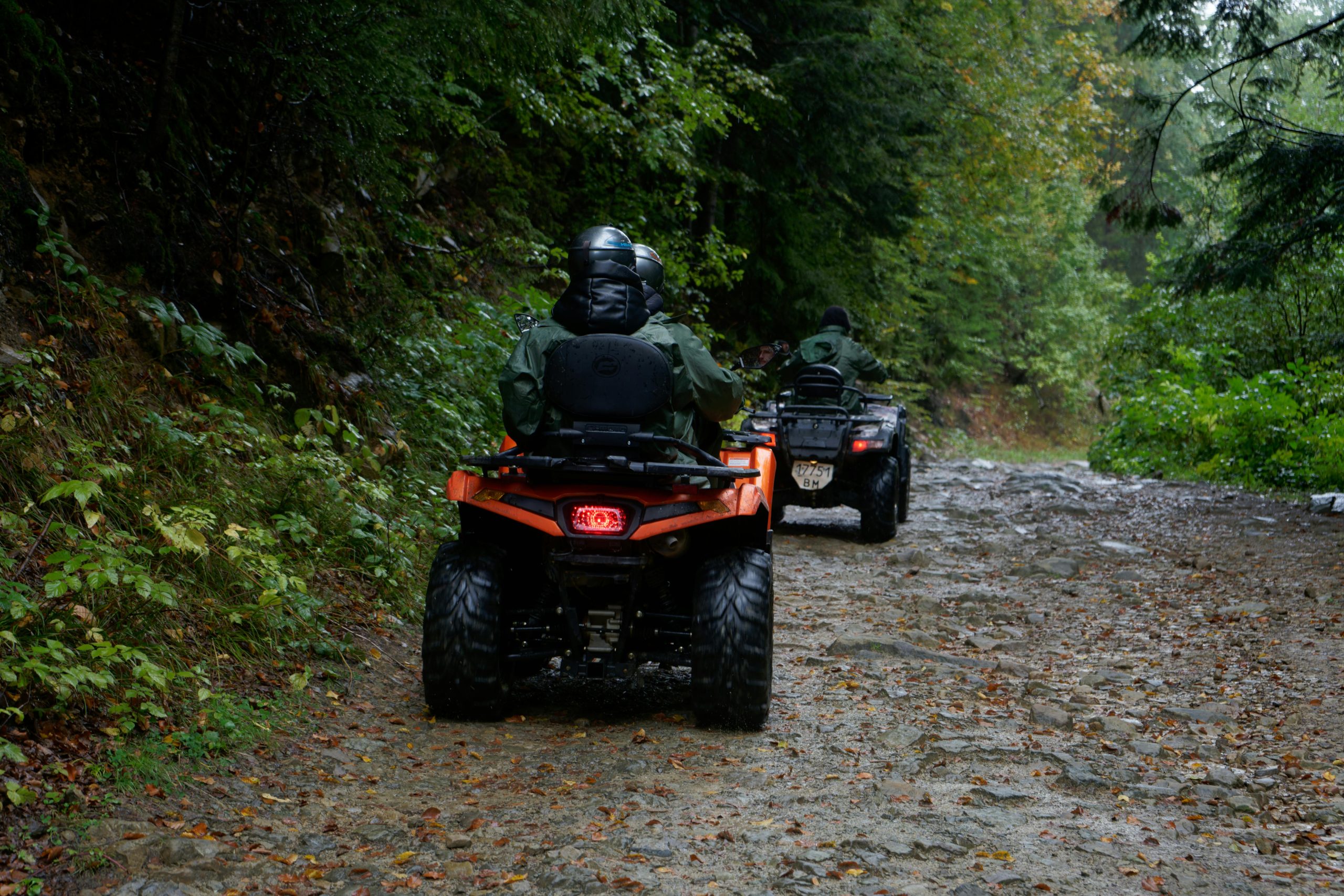 Two people riding ATVs on a forest dirt road, surrounded by lush greenery.